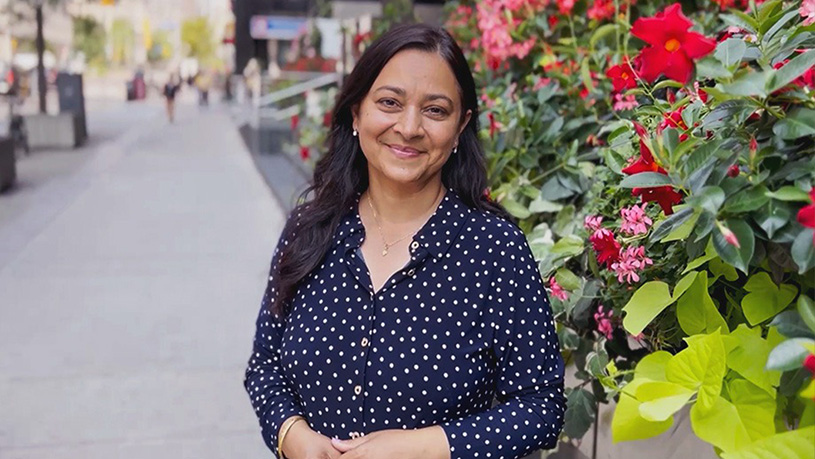 A woman stands on a city sidewalk beside a wall of bright red and pink flowers.