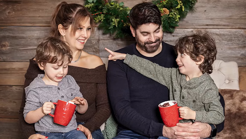 A family sits together holding red mugs in a cozy holiday setting with a wreath in the background.