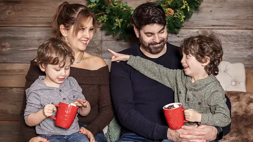 A family sits together holding red mugs in a cozy holiday setting with a wreath in the background.