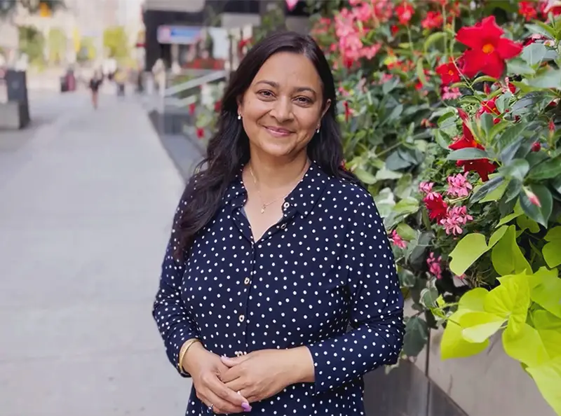 A woman stands on a city sidewalk beside a wall of bright red and pink flowers.